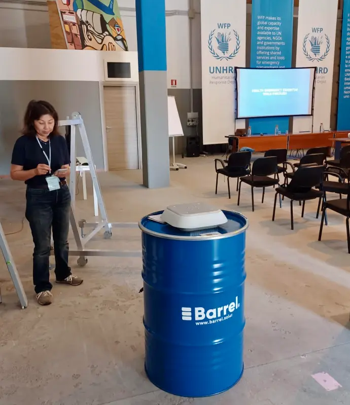 alt="Barrel solar energy system displayed at a UNHRD event with a participant and presentation setup in the background"