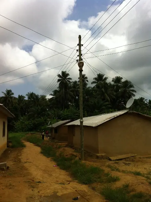 alt="Electric poles and wires in a rural village setting in Ghana, surrounded by palm trees"