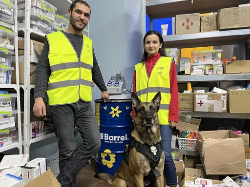 alt="Two volunteers and a rescue dog in a medical supply storage with a Barrel solar energy system in Ukraine"